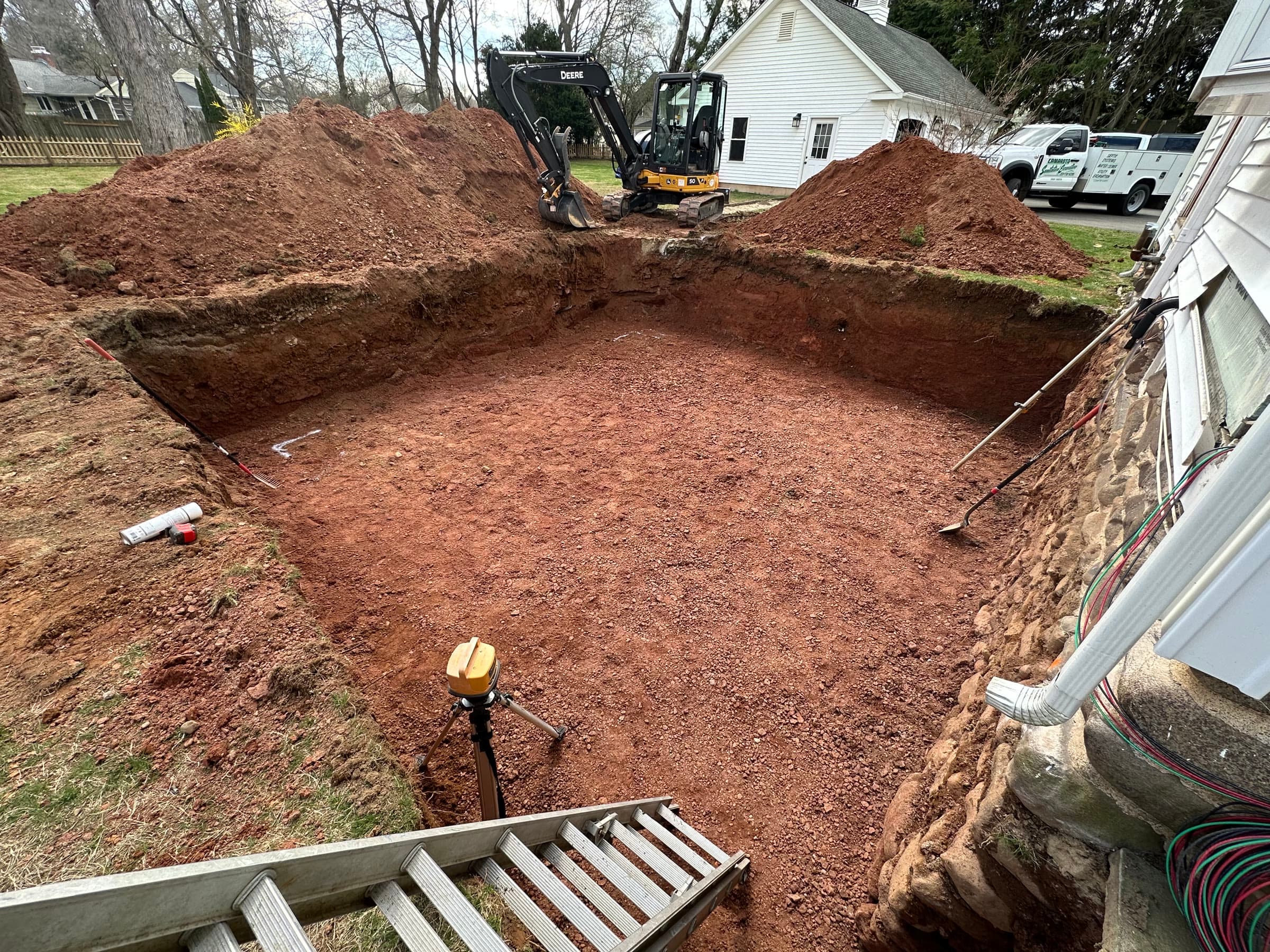 Foundation excavation: a rectangular cut of red soil beside a home, with a compact excavator on the spoil pile.