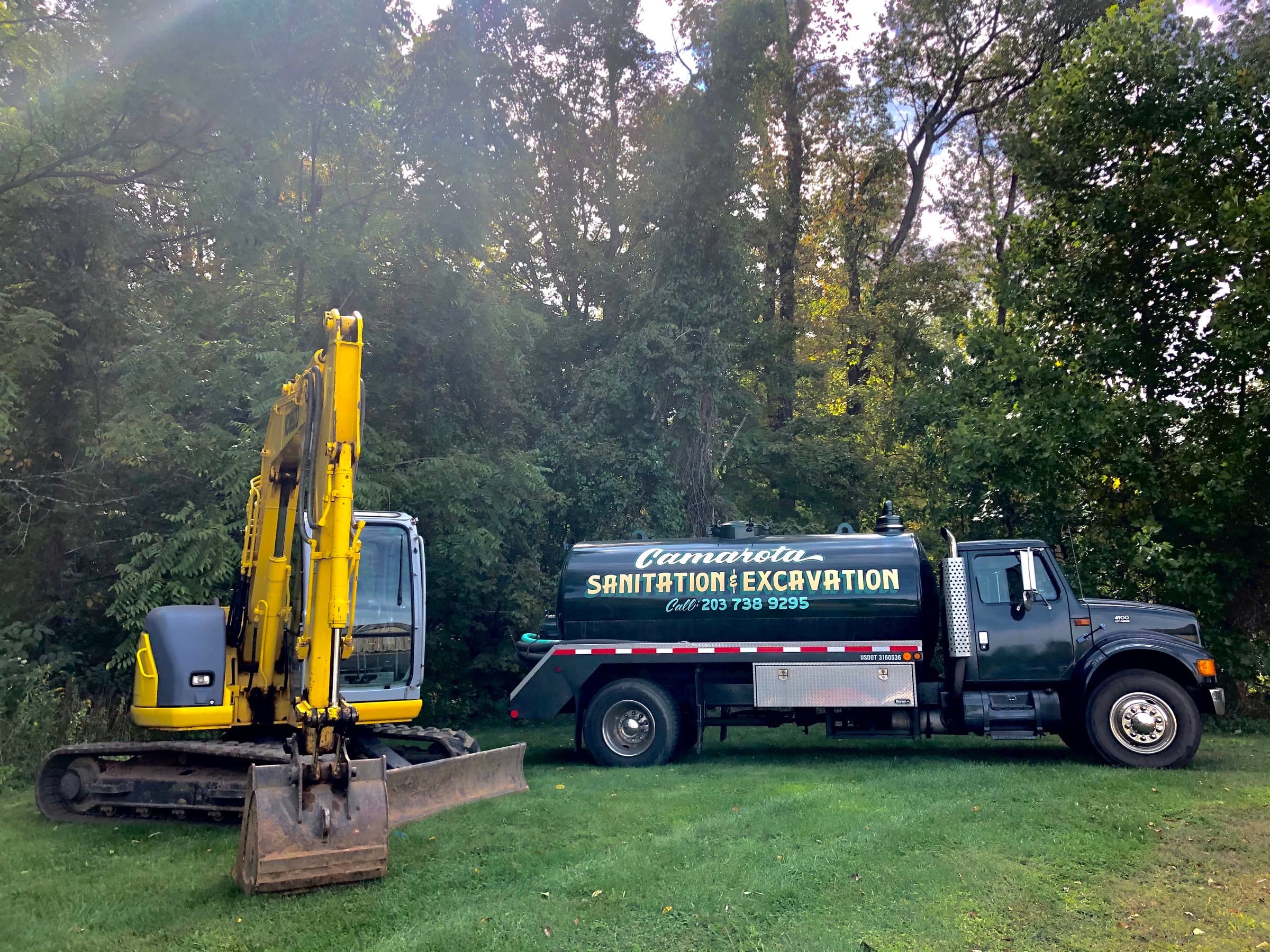 Camarota Sanitation & Excavation pump truck parked beside a yellow excavator on a grassy Shoreline lot.